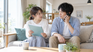 自宅のソファで書類を持ち、電話で家族の健康保険について確認する若い夫婦